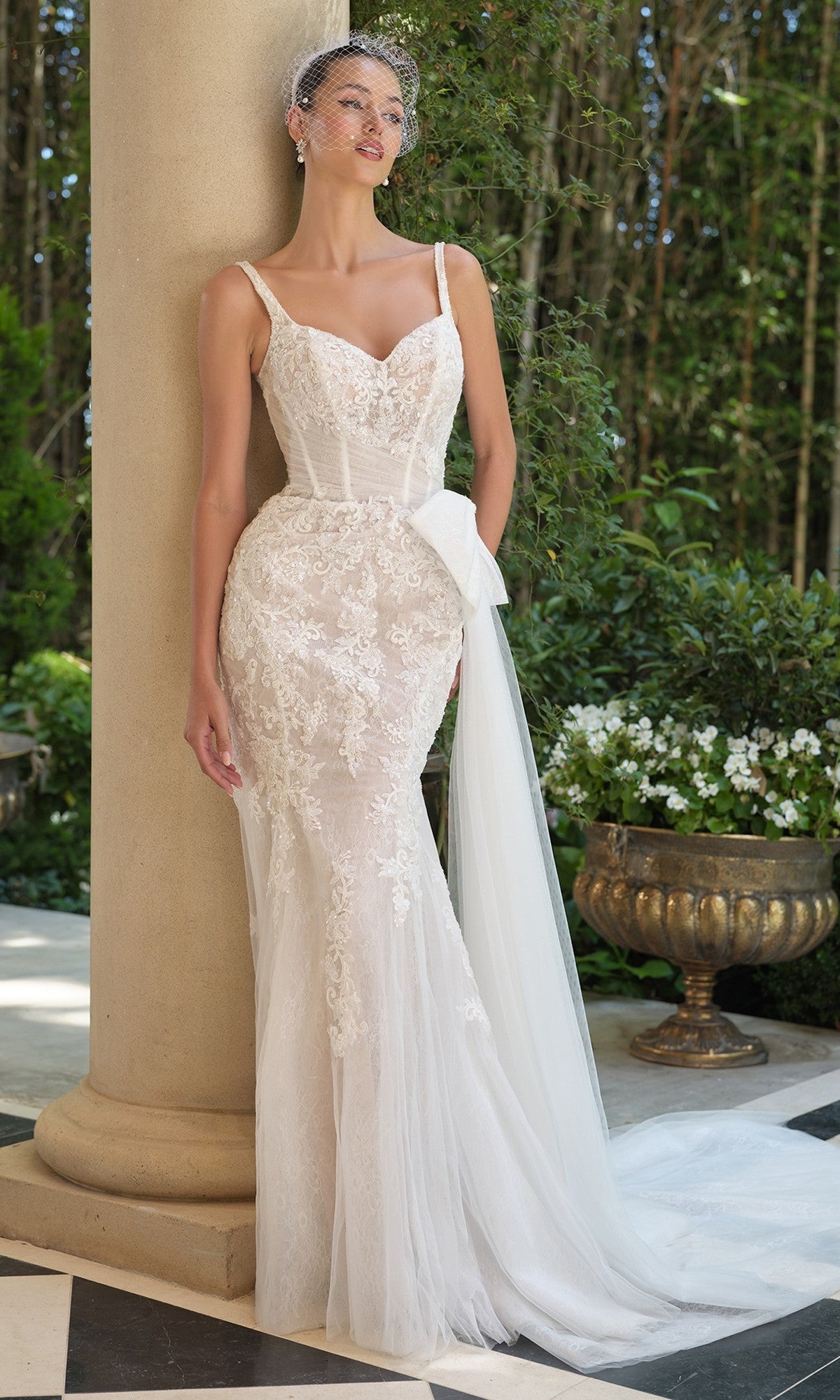 A woman in the Andrea and Leo WL068 long wedding dress with a lace design, sweetheart neckline, sheer corset bodice, and bow detail stands by a column in a tranquil garden.